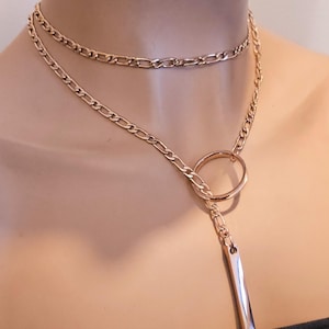 May include: A rose gold-toned layered necklace featuring a short figaro chain choker and a longer chain that passes through a circular ring, ending in a vertical rectangular bar pendant. The metal has a polished, reflective finish throughout.