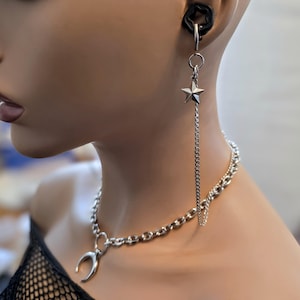 May include: A silver-coloured necklace with a crescent moon pendant and a matching chain. A star-shaped earring hangs from a chain. A black earring is also visible. The jewellery is displayed on a mannequin.