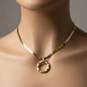 Peut inclure: Un collier doré avec une chaîne en chevrons et un pendentif circulaire au design ondulé texturé. Le collier est présenté sur un mannequin, mettant en valeur son style et son design élégants.