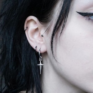 May include: Close-up of an ear with multiple silver earrings. A hoop, a small stud, and a dangling cross earring are visible. The person has dark hair and is wearing eyeliner.