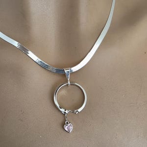 May include: A silver-toned herringbone chain necklace featuring a circular pendant. The pendant is attached to the chain by a bail and includes a small, dangling heart-shaped charm with a light pink stone set in the center of the heart.