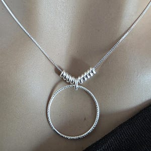 May include: A silver-toned necklace featuring a thin box chain and a circular pendant with a textured, twisted rope-like surface. The pendant is suspended from the chain by a series of five small, smooth, cylindrical silver-toned spacer beads.