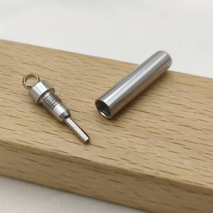 May include: A silver-toned metal cremation urn pendant consisting of two parts: a hollow cylindrical outer casing and a threaded inner plug with a small circular loop at the top for attachment to a chain or cord, shown disassembled.