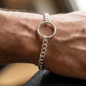May include: A silver-toned bracelet featuring a polished circular ring connector joined to two segments of a textured curb chain. The metallic links have a reflective finish, creating a continuous loop design that rests against the wrist.