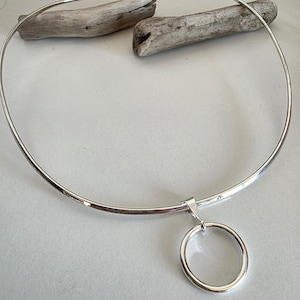 May include: A silver-toned metal collar necklace featuring a smooth, rigid circular band that rests in a wide arc. A single, polished silver-toned ring pendant hangs from a small triangular bail attached to the center of the collar necklace.