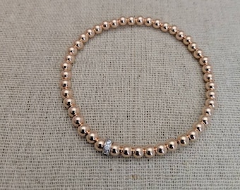 May include: Two beaded bracelets. The lower bracelet is rose gold with small round beads and a silver accent with clear stones. The upper bracelet is gold with a similar silver accent. Both bracelets are on a tan fabric surface.