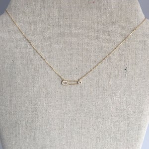 May include: A gold chain necklace with a small, diamond-encrusted safety pin pendant.