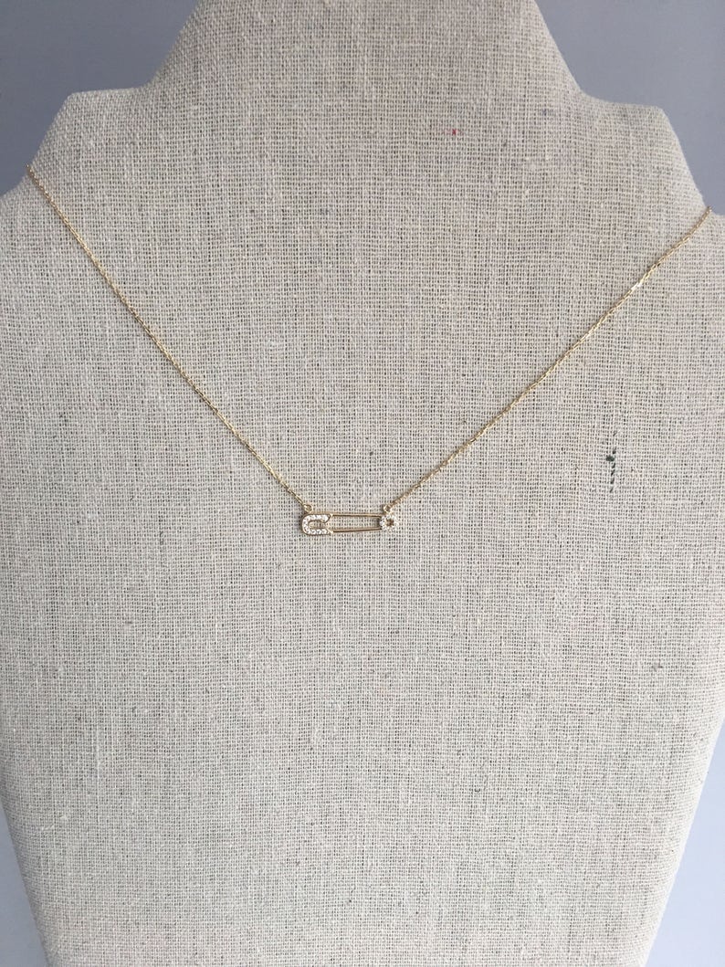 May include: A gold chain necklace with a small, diamond-encrusted safety pin pendant.