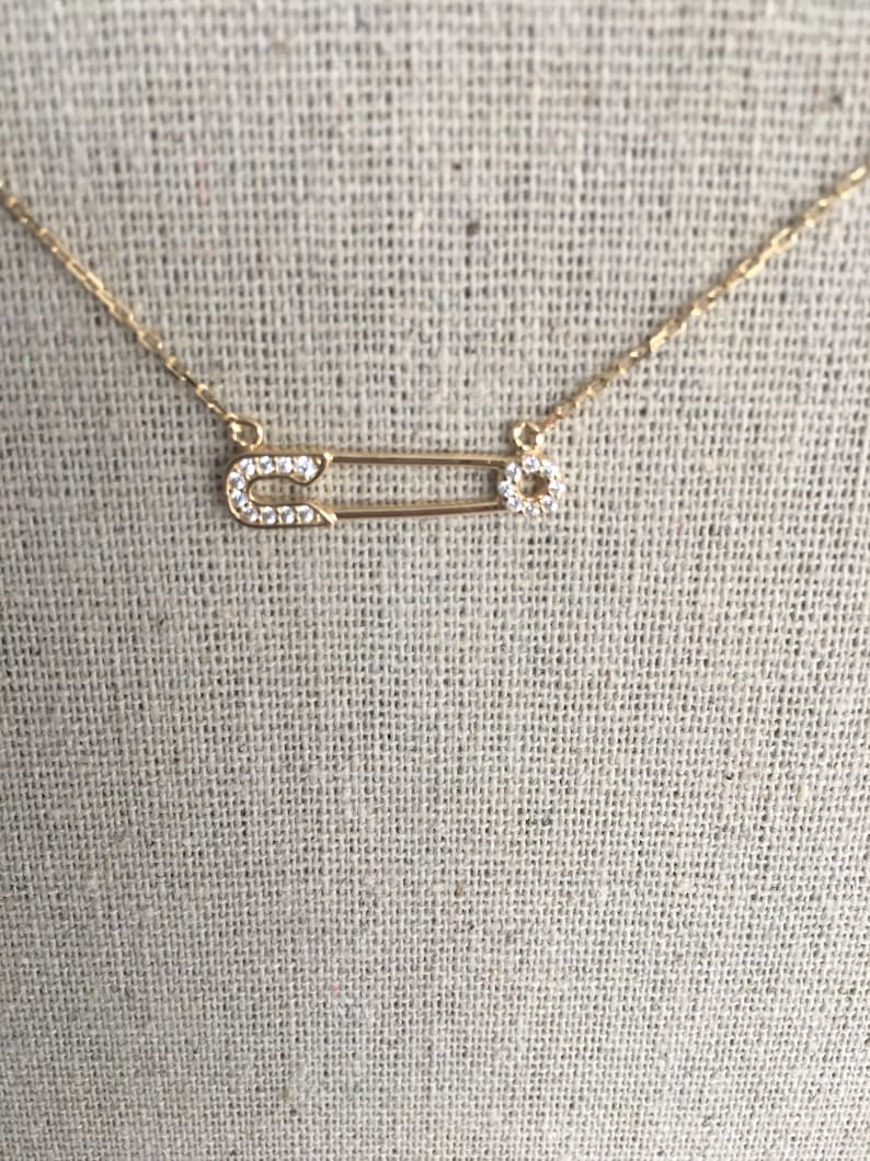 May include: A gold necklace with a safety pin pendant encrusted with clear rhinestones.
