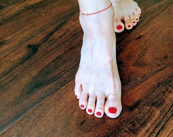 May include: A close-up of a person's bare feet with red nail polish. The person is wearing a red string bracelet with gold beads around their ankle.