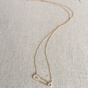 May include: A gold chain necklace with a small, diamond-encrusted safety pin pendant.