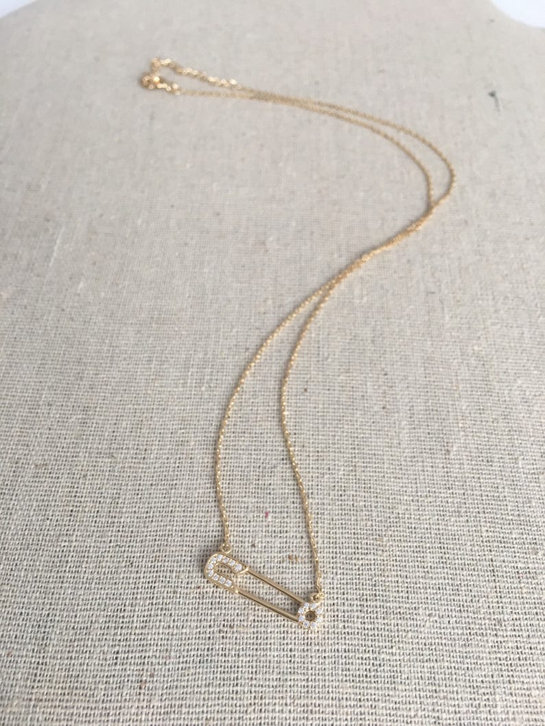 May include: A gold chain necklace with a small, diamond-encrusted safety pin pendant.