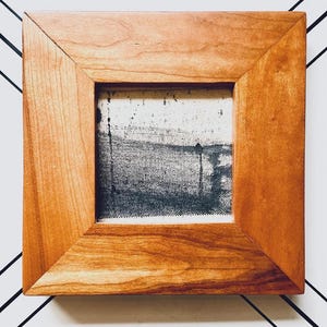 Puede incluir: Obra de arte cuadrada enmarcada con un marco de madera marrón claro. La obra de arte interior es una pintura de paisaje abstracta en blanco y negro. La pintura representa una escena con una línea de horizonte, posiblemente un cuerpo de agua, y elementos verticales.