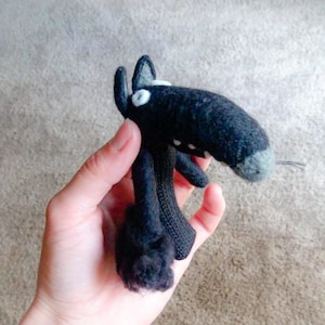 May include: A black knitted stuffed animal shaped like a wolf with white eyes. The wolf has a long snout and a small tail.