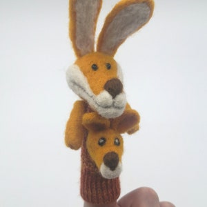May include: A finger puppet with two yellow felt rabbits stacked on top of each other. The top rabbit has white felt ears and a brown felt nose. The bottom rabbit has a brown felt nose and is wearing a red and orange knitted finger puppet.
