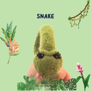 Könnte beinhalten: Eine grüne Filzschlangenpuppe mit schwarzen Wimpern und dem Wort "SNAKE" in blauen Buchstaben darüber. Die Puppe ist von grünen Blättern und Blumen umgeben.