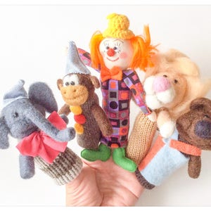 May include: Five finger puppets, including a clown with a yellow hat and orange hair, a monkey with a blue hat, an elephant with a blue and white striped hat, a lion with a light blue shirt, and a brown bear with a blue and orange shirt.