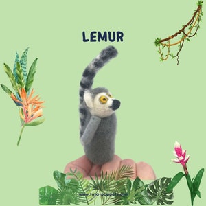 Peut inclure: Une marionnette de doigt en gris et blanc représentant un lémurien avec des anneaux noirs sur sa queue. Le lémurien lève les bras et a une expression surprise sur son visage. La marionnette est sur une main sur un fond vert avec des plantes tropicales.