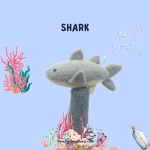 May include: A gray felt shark finger puppet with a white belly, surrounded by coral and bubbles. The text "SHARK" is above the puppet. The website "www.fato-puppets.com" is below the puppet.