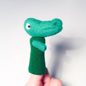 May include: Green felt finger puppet shaped like an alligator with a white eye. The alligator is wearing a green knitted sweater.