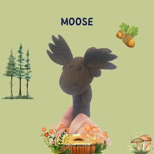 May include: A brown felt moose puppet with large antlers. The puppet is held by a hand against a green background with a small forest scene, mushrooms, and acorns.