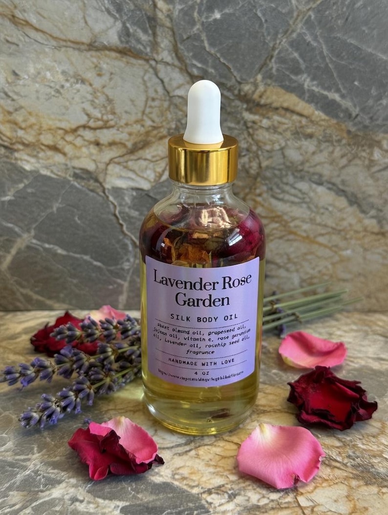 Puede incluir: Una botella de vidrio transparente de Lavender Rose Garden Silk Body Oil con tapa dorada y cuentagotas blanco. La etiqueta enumera los ingredientes y dice "HANDMADE WITH LOVE". Lavanda seca y p&eacute;talos de rosa rodean la botella. La botella contiene 118 ml de aceite.