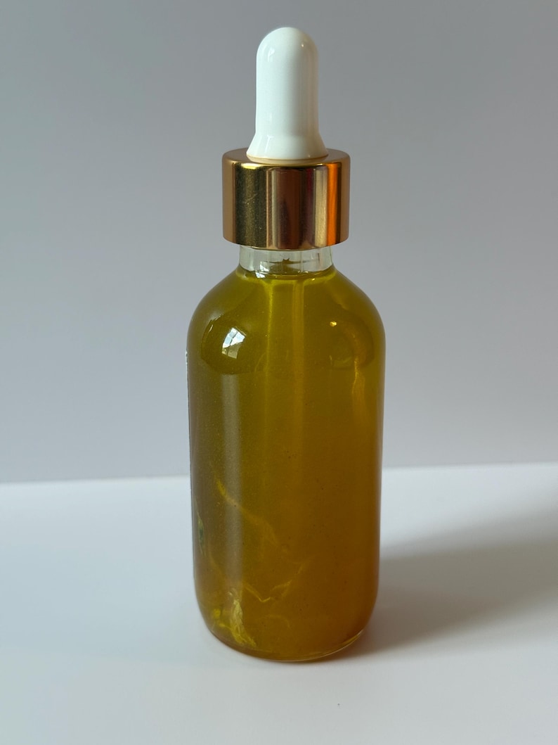 Puede incluir: Una botella de vidrio transparente llena de l&iacute;quido amarillo. Tiene una tapa dorada y un cuentagotas blanco. La botella cil&iacute;ndrica est&aacute; sobre una superficie blanca.