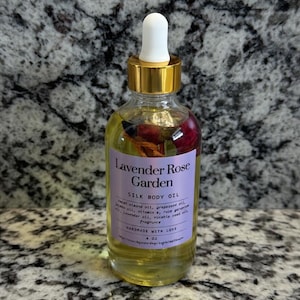 Puede incluir: Una botella de vidrio transparente de Lavender Rose Garden Silk Body Oil con tapa dorada y cuentagotas blanco. La etiqueta es morada con texto blanco. La botella contiene flores secas y es de 118 ml.