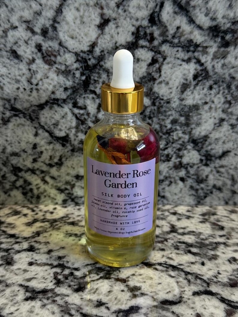 Puede incluir: Una botella de vidrio transparente de Lavender Rose Garden Silk Body Oil con tapa dorada y cuentagotas blanco. La etiqueta es morada con texto blanco. La botella contiene flores secas y es de 118 ml.