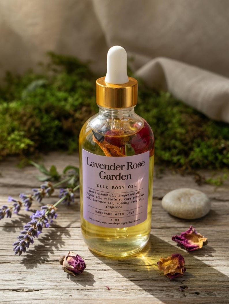 Puede incluir: Una botella de vidrio transparente de Lavender Rose Garden Silk Body Oil con tapa y cuentagotas dorados. La etiqueta enumera ingredientes como aceite de almendras dulces y aceite de lavanda. P&eacute;talos de rosa secos y ramitas de lavanda est&aacute;n dispuestos alrededor de la botella. La botella contiene 118 ml de aceite.