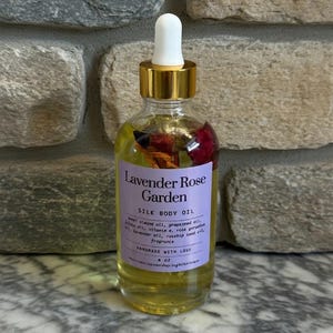 Puede incluir: Una botella de vidrio transparente de Lavender Rose Garden Silk Body Oil con tapa dorada y cuentagotas blanco. La etiqueta es morada con texto blanco. La botella contiene flores secas y es de 118 ml.