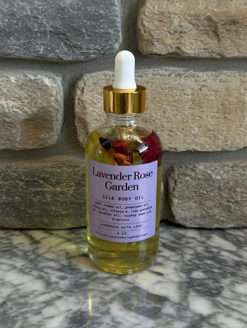 Puede incluir: Una botella de vidrio transparente de Lavender Rose Garden Silk Body Oil con tapa dorada y cuentagotas blanco. La etiqueta es morada con texto blanco. La botella contiene flores secas y es de 118 ml.