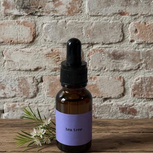 May include: A brown glass bottle with a black dropper top and a purple label that reads "tea tree." The bottle sits on a wooden surface next to a sprig of white flowers and green leaves. The background is a brick wall.