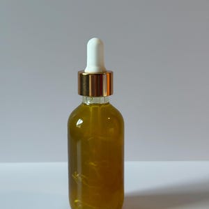 Puede incluir: Una botella de vidrio transparente llena de un l&iacute;quido dorado. La botella tiene un cuentagotas blanco y un collar de metal dorado. Se encuentra sobre una superficie blanca.