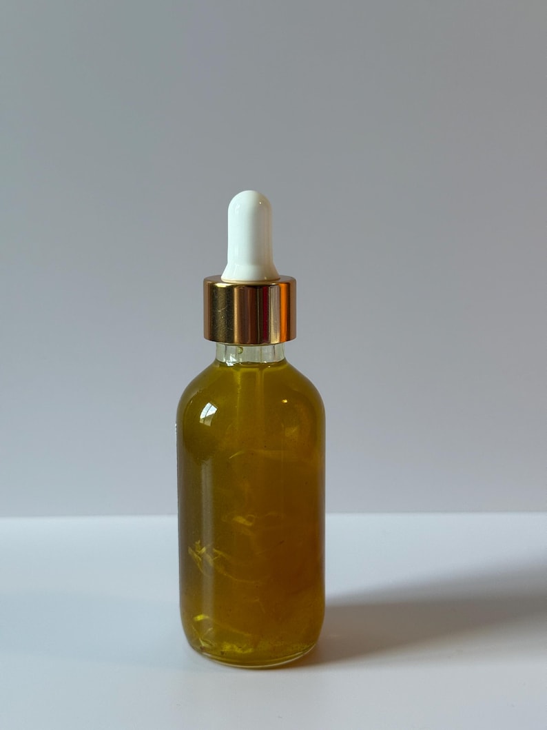 Puede incluir: Una botella de vidrio transparente llena de un l&iacute;quido dorado. La botella tiene un cuentagotas blanco y un collar de metal dorado. Se encuentra sobre una superficie blanca.