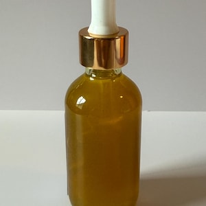 Puede incluir: Una botella de vidrio transparente llena de un l&iacute;quido dorado. Tiene una tapa de metal dorado y un cuentagotas blanco. La botella est&aacute; sobre una superficie blanca.