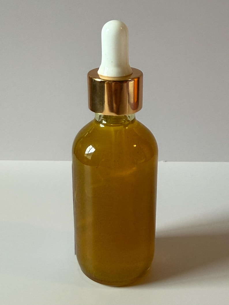 Puede incluir: Una botella de vidrio transparente llena de un l&iacute;quido dorado. Tiene una tapa de metal dorado y un cuentagotas blanco. La botella est&aacute; sobre una superficie blanca.