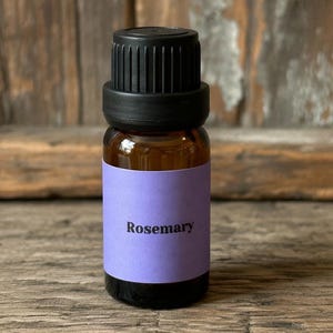 May include: A small, brown glass bottle with a black cap and a purple label. The label reads "Rosemary" in a simple, sans-serif font. The bottle is likely filled with essential oil, and is displayed on a wooden surface.