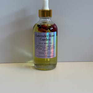 Puede incluir: Una botella de vidrio transparente de Lavender Rose Garden Silk Body Oil con tapa dorada y cuentagotas blanco. La etiqueta incluye el nombre del producto y los ingredientes. La botella contiene flores secas y es de 118 ml.