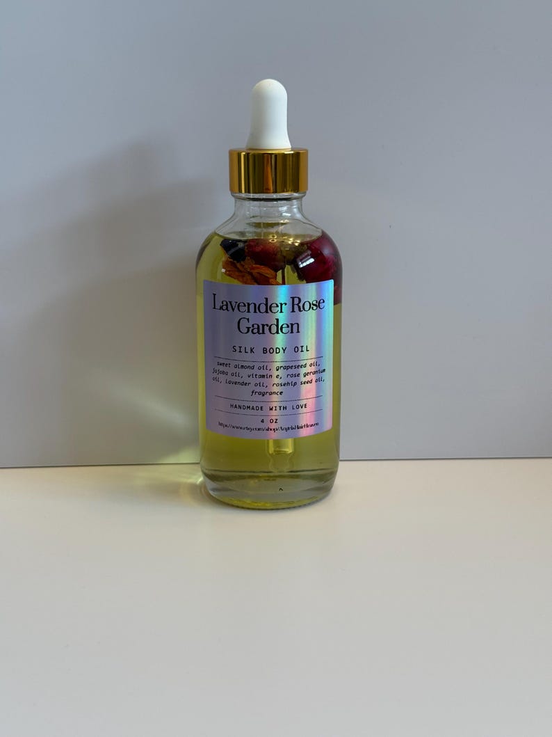 Puede incluir: Una botella de vidrio transparente de Lavender Rose Garden Silk Body Oil con tapa dorada y cuentagotas blanco. La etiqueta incluye el nombre del producto y los ingredientes. La botella contiene flores secas y es de 118 ml.
