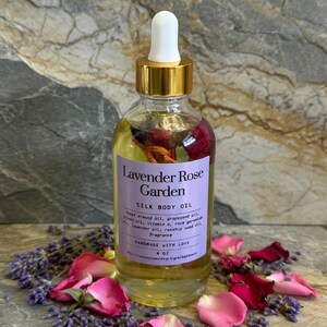 Puede incluir: Una botella de vidrio transparente de Lavender Rose Garden Silk Body Oil con tapa dorada y cuentagotas blanco. La etiqueta enumera los ingredientes. La botella est&aacute; rodeada de p&eacute;talos de rosa rosas y capullos de lavanda. La botella contiene 118 ml de aceite.