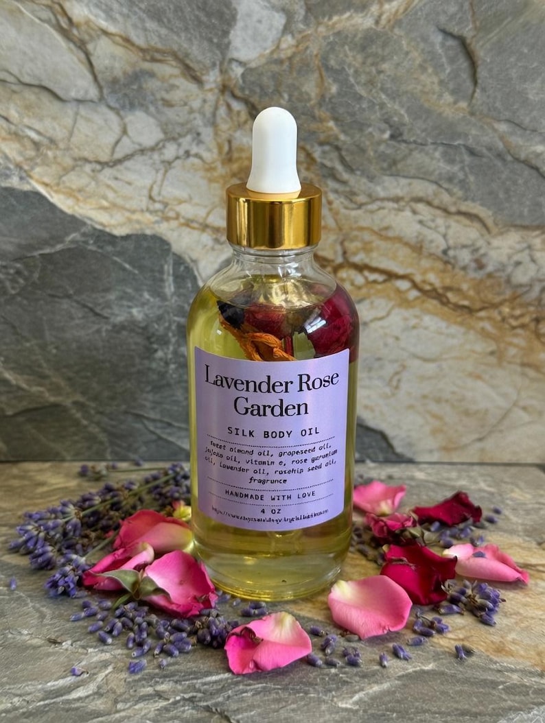 Puede incluir: Una botella de vidrio transparente de Lavender Rose Garden Silk Body Oil con tapa dorada y cuentagotas blanco. La etiqueta enumera los ingredientes. La botella est&aacute; rodeada de p&eacute;talos de rosa rosas y capullos de lavanda. La botella contiene 118 ml de aceite.