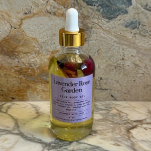 Puede incluir: Una botella de vidrio transparente de Lavender Rose Garden Silk Body Oil con una tapa cuentagotas blanca y un collar dorado. La etiqueta es morada con texto blanco. La botella contiene p&eacute;talos de rosa secos. La botella es de 118 ml.