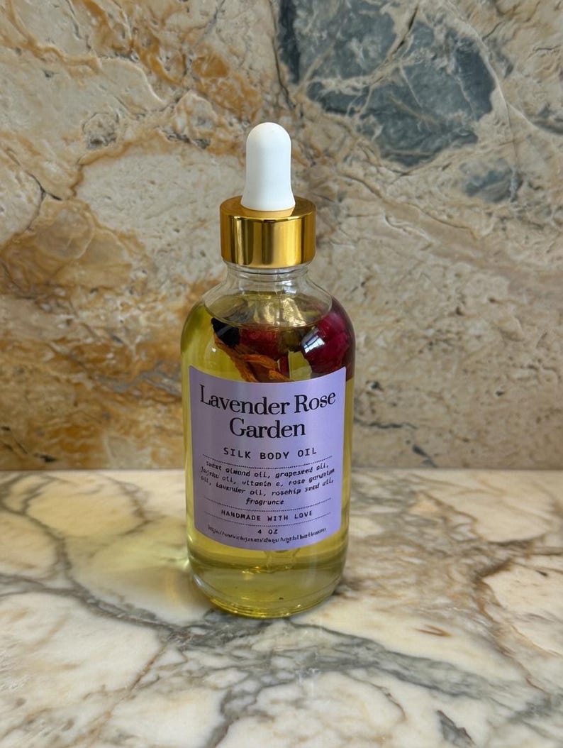 Puede incluir: Una botella de vidrio transparente de Lavender Rose Garden Silk Body Oil con una tapa cuentagotas blanca y un collar dorado. La etiqueta es morada con texto blanco. La botella contiene p&eacute;talos de rosa secos. La botella es de 118 ml.