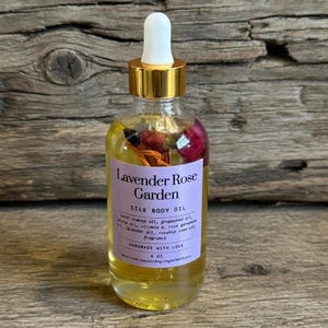 Puede incluir: Una botella de vidrio transparente de aceite corporal de seda Lavender Rose Garden con tapa dorada y cuentagotas blanco. La botella contiene flores secas y aceite. La etiqueta dice "Lavender Rose Garden". La botella es de 118 ml.
