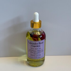 Puede incluir: Una botella de vidrio transparente de aceite corporal de seda Lavender Rose Garden con tapa dorada y cuentagotas blanco. La botella contiene un l&iacute;quido amarillo con flores secas. La etiqueta es morada con texto blanco.