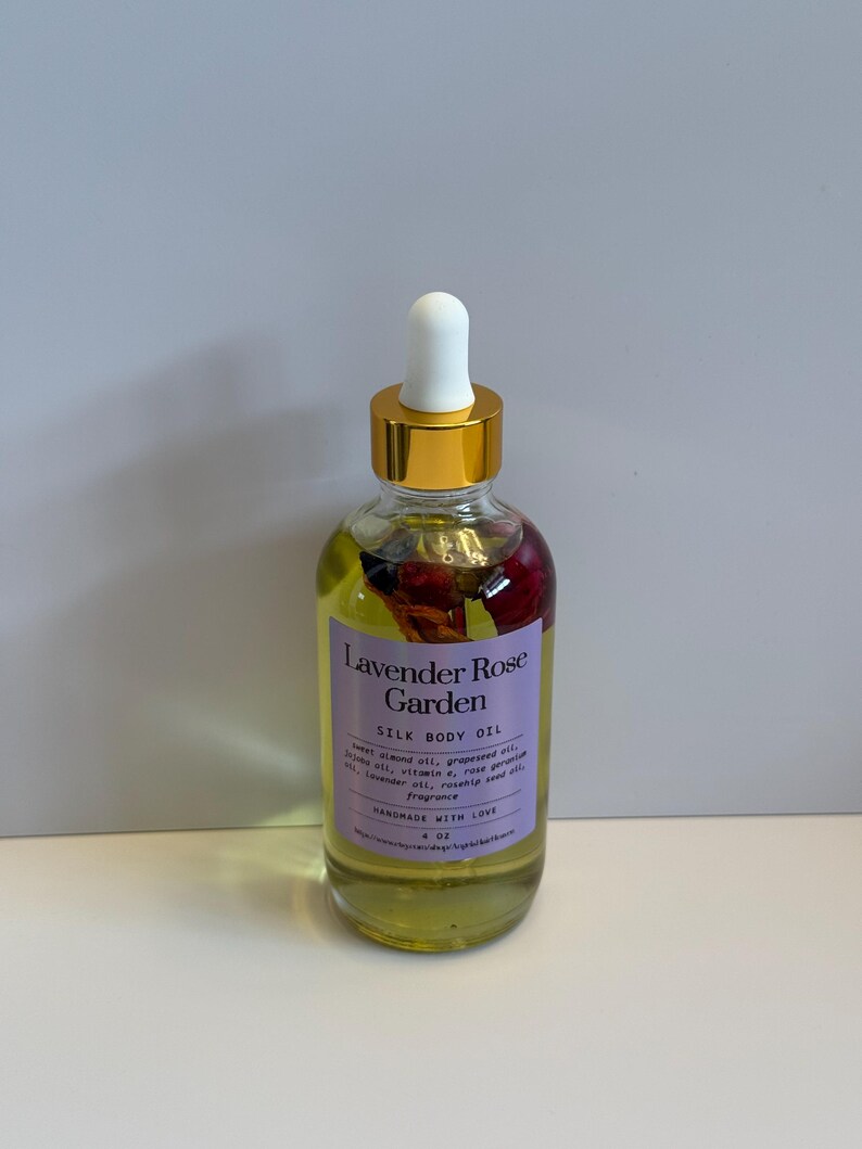 Puede incluir: Una botella de vidrio transparente de aceite corporal de seda Lavender Rose Garden con tapa dorada y cuentagotas blanco. La botella contiene un l&iacute;quido amarillo con flores secas. La etiqueta es morada con texto blanco.