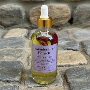 Puede incluir: Una botella de vidrio transparente de Lavender Rose Garden Silk Body Oil con tapa y cuentagotas dorados. La etiqueta es morada con texto blanco. La botella contiene flores secas y aceite. La botella es de 118 ml.