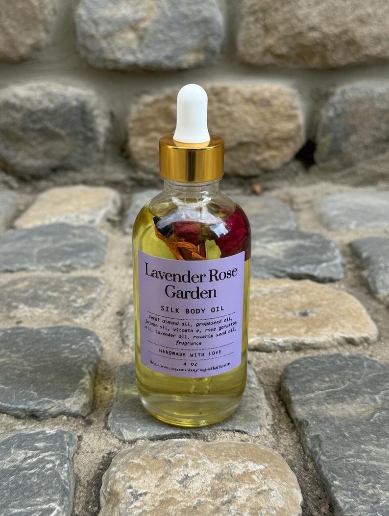 Puede incluir: Una botella de vidrio transparente de Lavender Rose Garden Silk Body Oil con tapa y cuentagotas dorados. La etiqueta es morada con texto blanco. La botella contiene flores secas y aceite. La botella es de 118 ml.