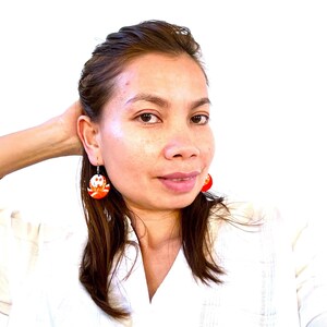 Puede incluir: Un par de pendientes circulares colgantes con un vibrante patrón abstracto en naranja y blanco. Los pendientes cuelgan de ganchos de metal plateado y muestran un diseño gráfico distintivo con líneas curvas y bloques de color.